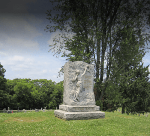 A stone monument in a cemetery featuring a figure with an outstretched arm, surrounded by trees and gravestones.