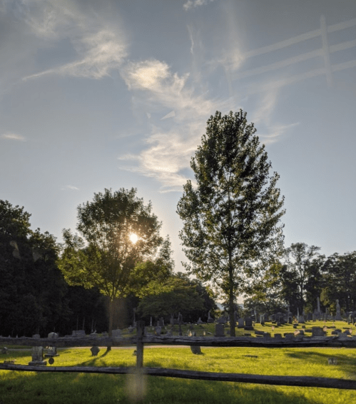 A serene cemetery scene with gravestones, trees, and a setting sun against a blue sky with wispy clouds.
