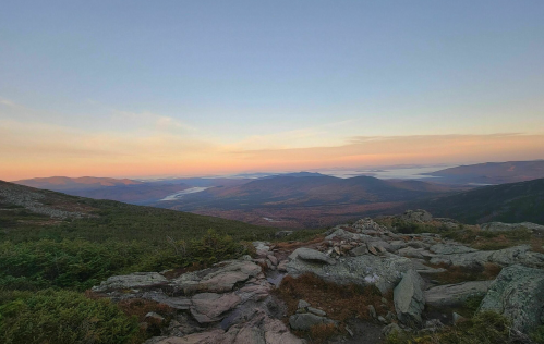 A panoramic view of mountains and valleys at sunset, with soft pastel colors in the sky and rocky foreground.