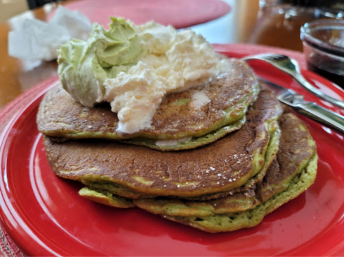 Three green pancakes stacked on a red plate, topped with whipped cream and a dollop of avocado.