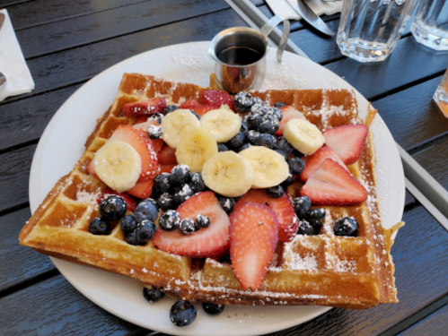 A plate of waffles topped with sliced bananas, strawberries, blueberries, and a small cup of syrup.