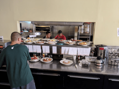 A busy kitchen with chefs preparing food behind a counter filled with plates of dishes and a coffee pot nearby.