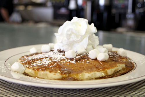 A stack of pancakes topped with whipped cream, caramel drizzle, and mini marshmallows on a white plate.
