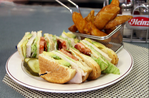 A plate with a club sandwich, layered with turkey, lettuce, tomato, and pickles, alongside a basket of golden fries.