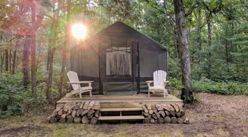 A cozy tent cabin with two white chairs on a wooden deck, surrounded by trees and sunlight filtering through.