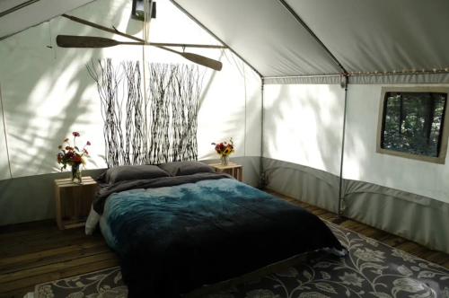 Cozy glamping tent interior with a bed, decorative wall, and flowers, surrounded by natural light and wooden flooring.