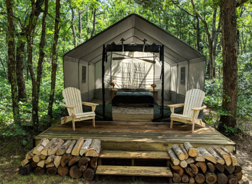 A cozy tent on a wooden deck in a forest, featuring two chairs and stacked logs in front.