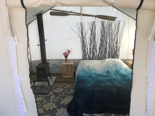 Cozy tent interior with a bed, decorative branches, a wood stove, and wooden crates.