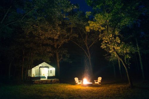 A cozy campsite at night with a glowing fire pit, two chairs, and a tent surrounded by trees.