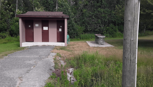 Two restroom buildings with a picnic table nearby, surrounded by grass and trees in a park setting.