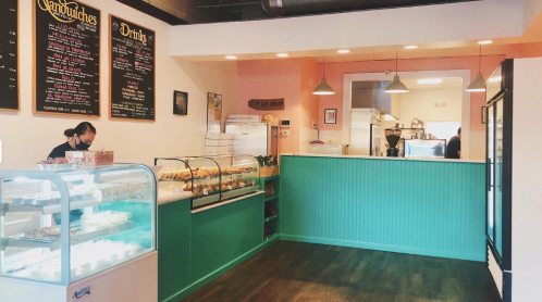 A cozy café interior with a display case, menu board, and staff member in a mask, featuring green and pink decor.