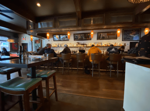 A cozy bar scene with patrons seated at the counter, warm lighting, and a well-stocked shelf of bottles in the background.