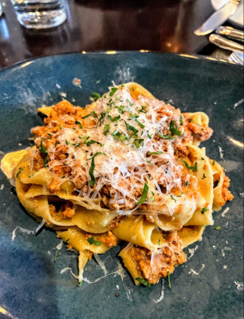 A plate of pappardelle pasta topped with a meat sauce and grated cheese, garnished with fresh herbs.