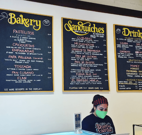 A menu board displaying bakery items, sandwiches, and drinks, with a staff member wearing a mask in front.