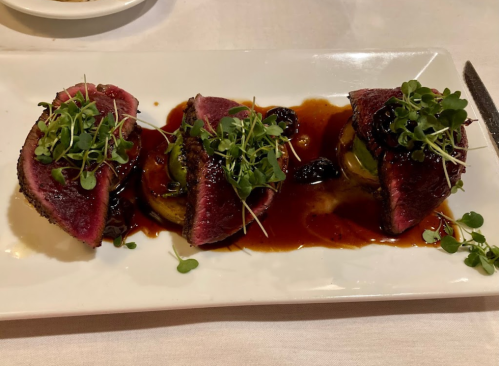 Three slices of medium-rare beef topped with microgreens, served on a plate with a rich sauce and garnishes.