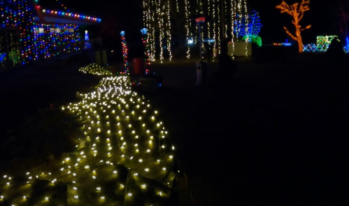 A pathway illuminated by warm white lights, surrounded by colorful holiday decorations in a dark setting.