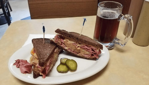 A plate with a stacked sandwich of corned beef on rye, pickles, and a glass of dark beer on the side.