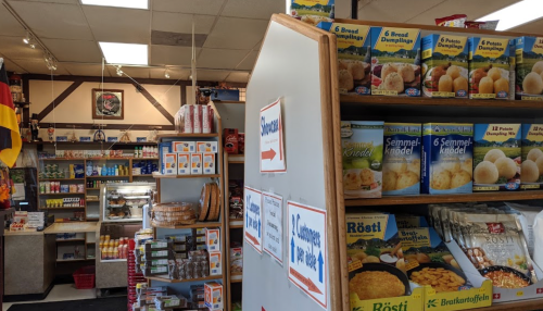 Interior of a small grocery store with shelves of packaged food items and signs directing customers.