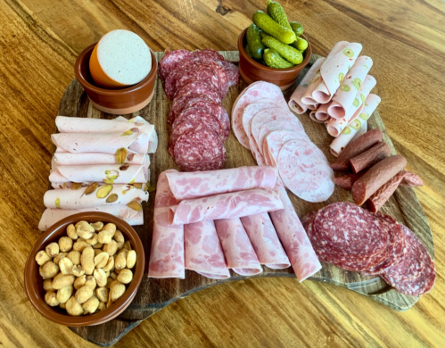 A wooden platter with various deli meats, cheeses, pickles, and peanuts arranged in small bowls.