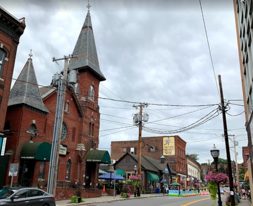 Historic buildings line a street with outdoor dining, power lines, and a cloudy sky overhead.