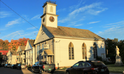 A white church with a clock tower, surrounded by trees and parked cars, under a blue sky with scattered clouds.