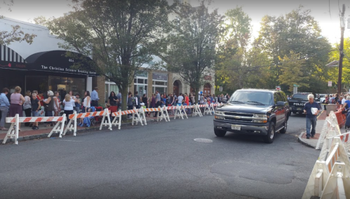 A line of people waiting outside a building, with police vehicles and barricades on a street. Trees line the background.