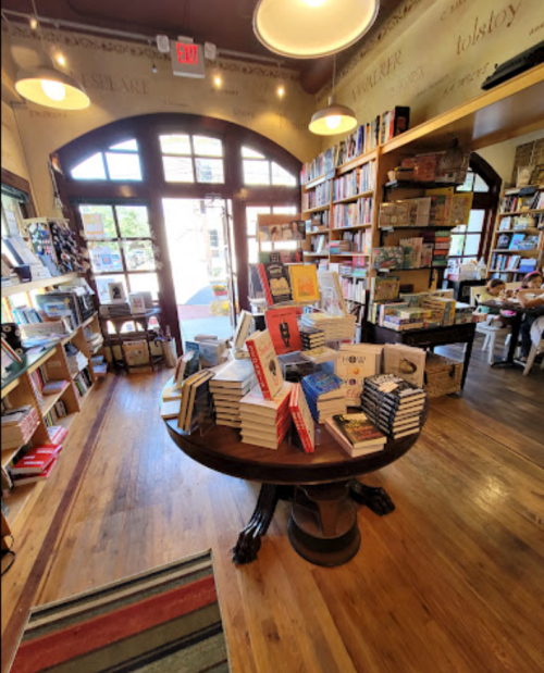 A cozy bookstore interior with shelves of books, a central table stacked with various titles, and large windows.