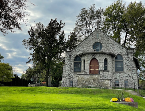 A stone church with arched windows, surrounded by green grass and trees under a cloudy sky.