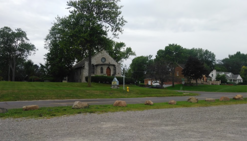 A stone building with a round window, surrounded by trees and houses, set against a cloudy sky. Gravel road in foreground.