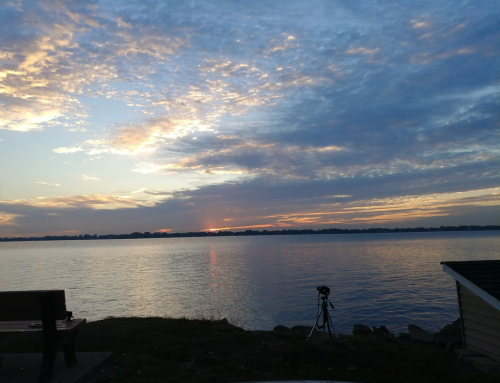 A serene lakeside view at sunset, with clouds reflecting colors in the water and a tripod set up nearby.