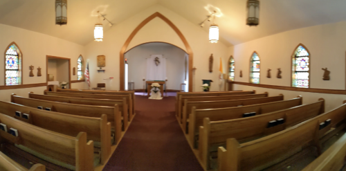Interior of a small church with wooden pews, stained glass windows, and a simple altar at the front.