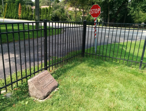 A stop sign at a corner near a black metal fence and a stone marker on a grassy area.