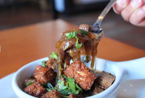 A spoonful of savory dish with crispy cubes, green onions, and a rich sauce being lifted from a bowl.