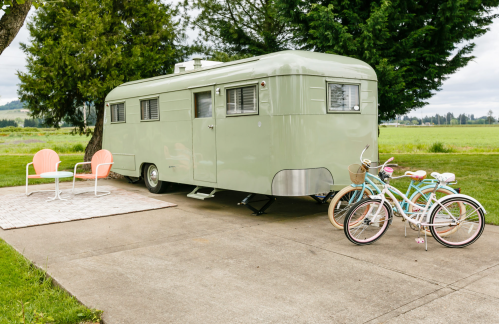 A vintage green trailer parked beside two bicycles and outdoor chairs on a concrete pad, surrounded by greenery.