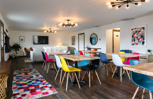Bright, modern communal space with colorful chairs, wooden tables, and a cozy seating area. Decor includes a rug and artwork.