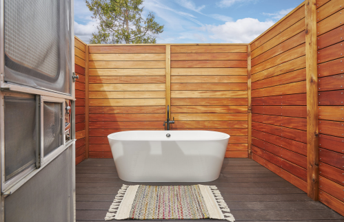 A modern outdoor bathroom featuring a freestanding white bathtub surrounded by wooden walls and a colorful rug.