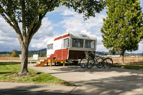A vintage camper parked on a concrete pad, surrounded by trees and two bicycles nearby, with a scenic landscape in the background.
