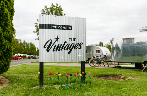 Sign reading "Welcome to The Vintages" with colorful flowers in front and vintage trailers in the background.