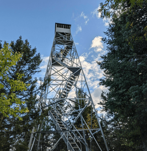 A tall metal fire lookout tower surrounded by trees under a blue sky with scattered clouds.