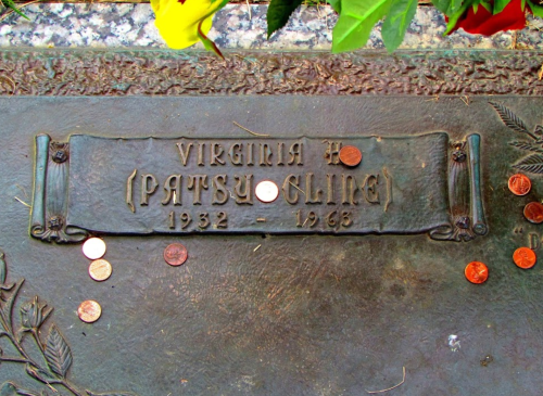 A bronze grave marker for Virginia H. (Patsy Cline) with coins placed on it, surrounded by flowers.