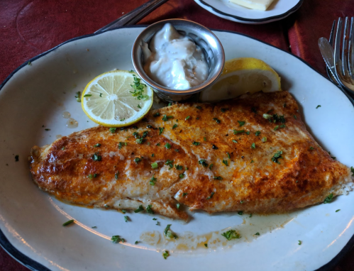A plated fish fillet garnished with parsley, lemon slices, and a small bowl of sauce on a red tablecloth.