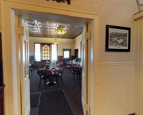 View through an open doorway into a dining room with tables set for a meal, featuring vintage decor and large windows.