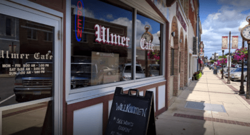 Exterior of Ulmer Cafe with an "Open" sign, sidewalk, and decorative banners in a small town setting.