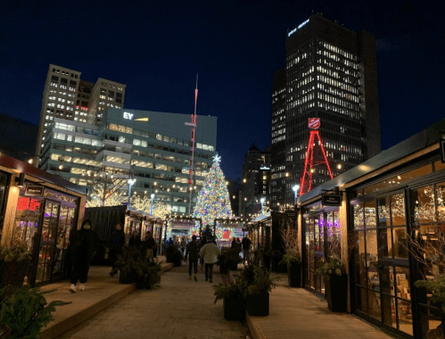 A festive market at night, featuring a large Christmas tree and illuminated buildings in a city setting.