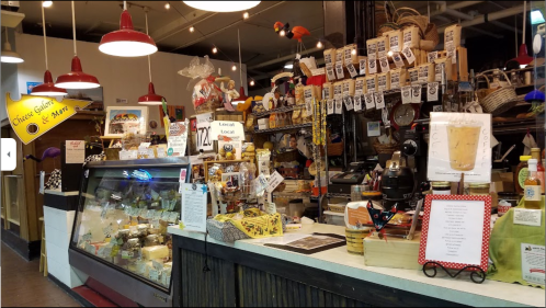 A cozy market display featuring various cheeses, baskets, and local products, with red pendant lights overhead.