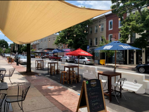 Outdoor dining area with tables and umbrellas, lined along a street with parked cars and trees in the background.