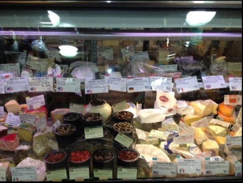 A display case filled with various cheeses, olives, and labeled products in a grocery store.