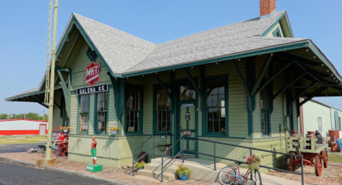 Historic train station with green siding, a peaked roof, and vintage signage, surrounded by old equipment and a clear blue sky.