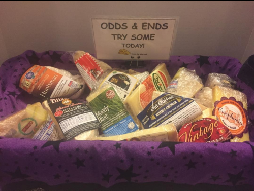 A basket filled with various cheese samples, labeled "Odds & Ends, Try Some Today!" on a purple cloth.