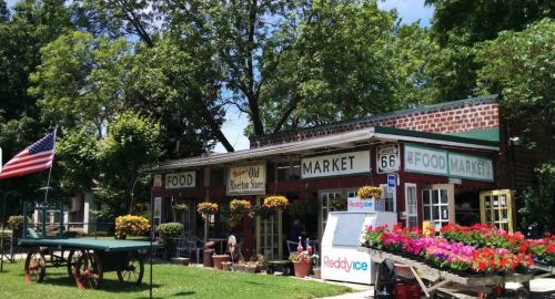 A charming market with a vintage sign, surrounded by flowers and greenery, featuring an American flag in front.